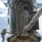 Two astronauts working on Hubble while in space. One astronaut is on the left, holding on to the lower handrail of Hubble. The astronaut on the right is standing on top of the CANADARM, a long robotic arm, he works on the center bottom of Hubble. In the distance behind Hubble, the blue Earth is visible with wisps of white clouds.