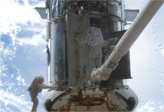 Image showing two astronauts working on Hubble while in space. One astronaut is on the left, holding on to the lower handrail of Hubble. The astronaut on the right is standing on top of the CANADARM, a long robotic arm, he works on the center bottom of Hubble. In the distance behind Hubble, the blue Earth is visible with wisps of white clouds.