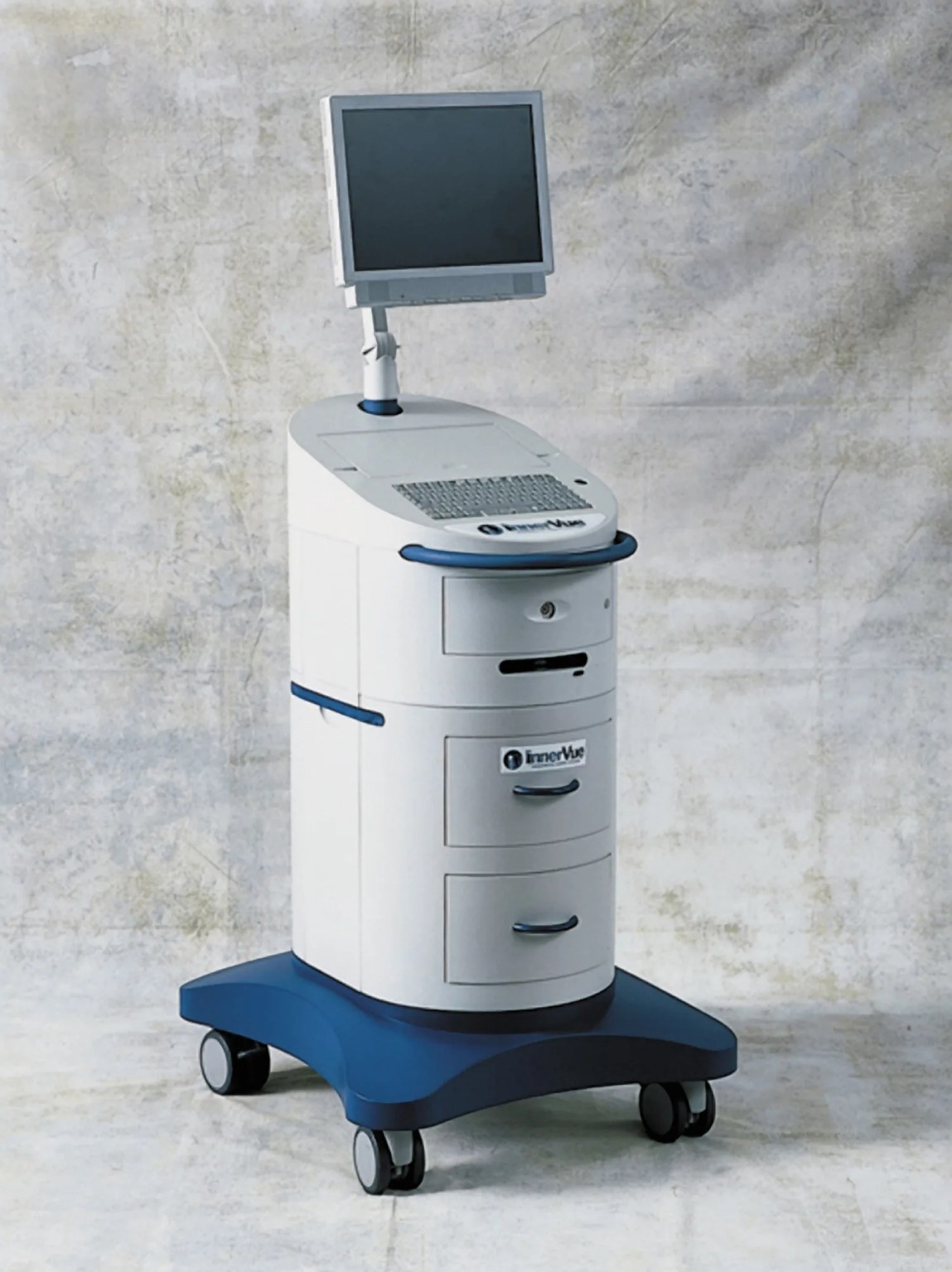 A roughly cyndrilical piece of equipment with three drawers and a handle for manuvering, a screen and keyboard on top, resting on a blue wheeled platform.