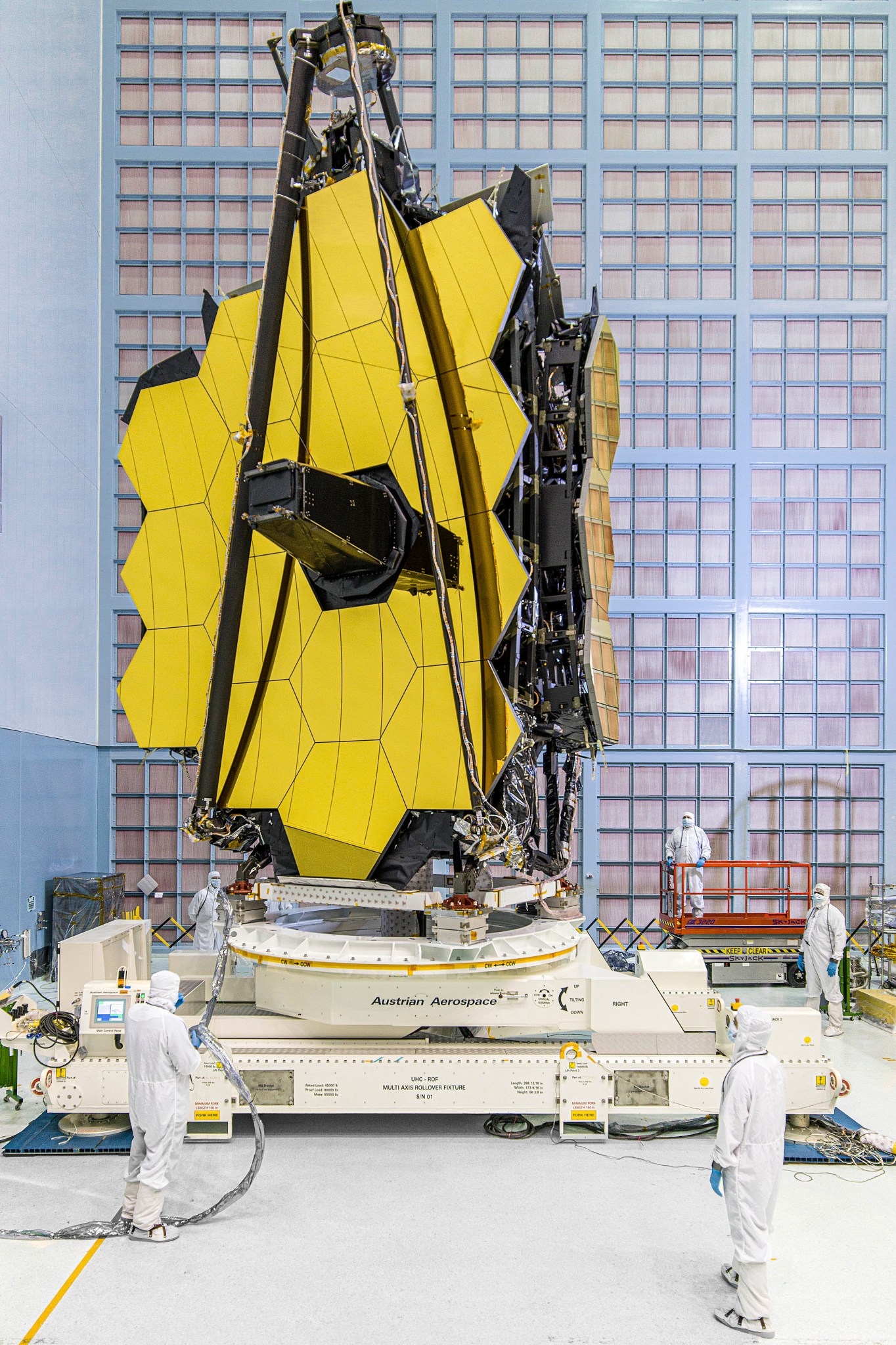 The James Webb Space Telescope mirrors upright on a white stand in the NASA Goddard cleanroom. The 3 mirrors on the right āwingā are folding back like a leaf on a drop-leaf table. The telescope has 18 golden hexagonal segments. The secondary mirror support structure, made of three thin black lines, is folded and the secondary mirror sits atop this folded tripod. There are 5 people in white cleanroom suits supervising the folding maneuver. One of them is on a red lift at the back right. There is a wall of HEPA filters behind the telescope.
