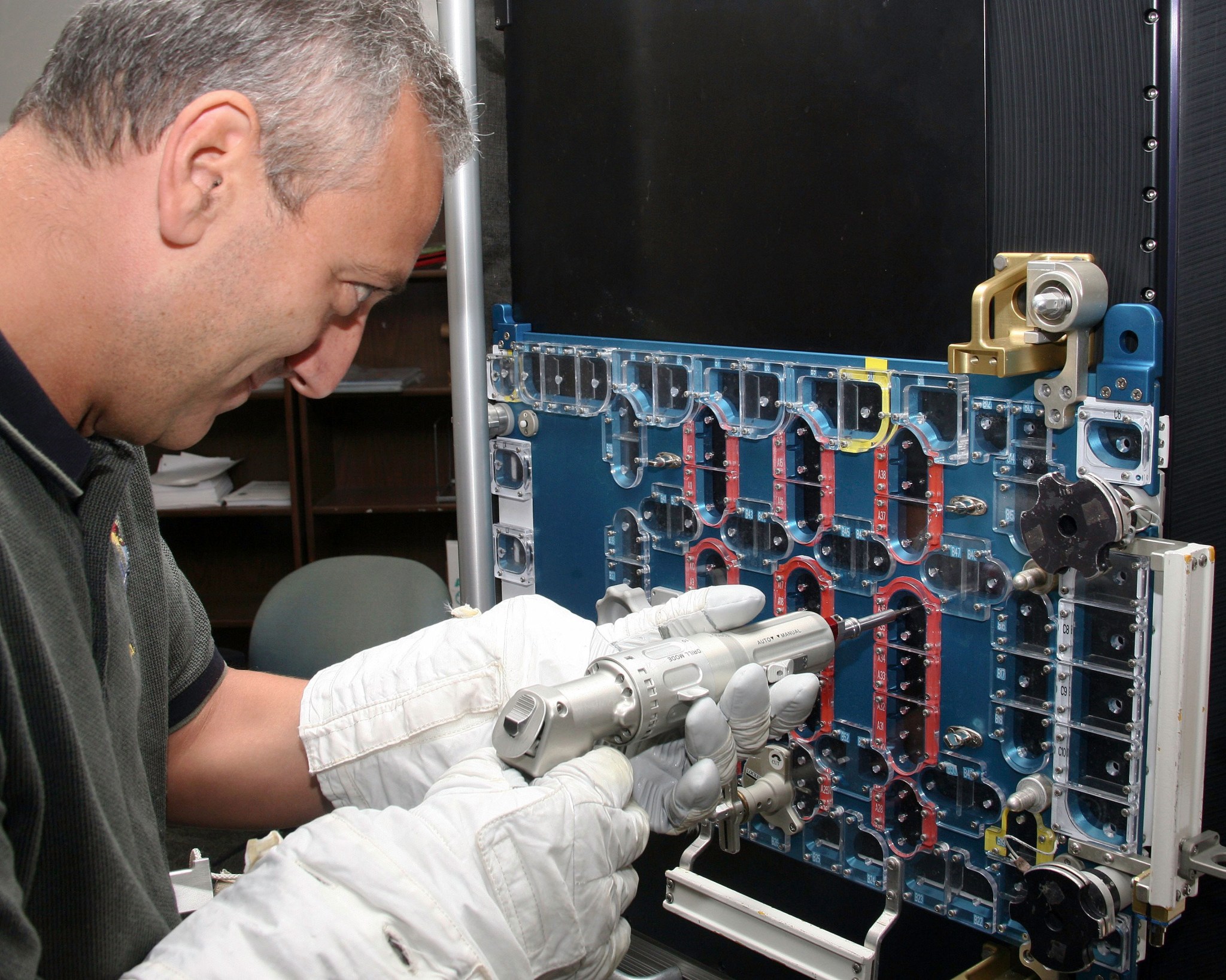 Wearing bulky white spacesuit gloves and a gray shirt, astronaut Mike Massimino holds a small, drill-shaped tool and uses it to remove screws through a flat blue device covered with holes.