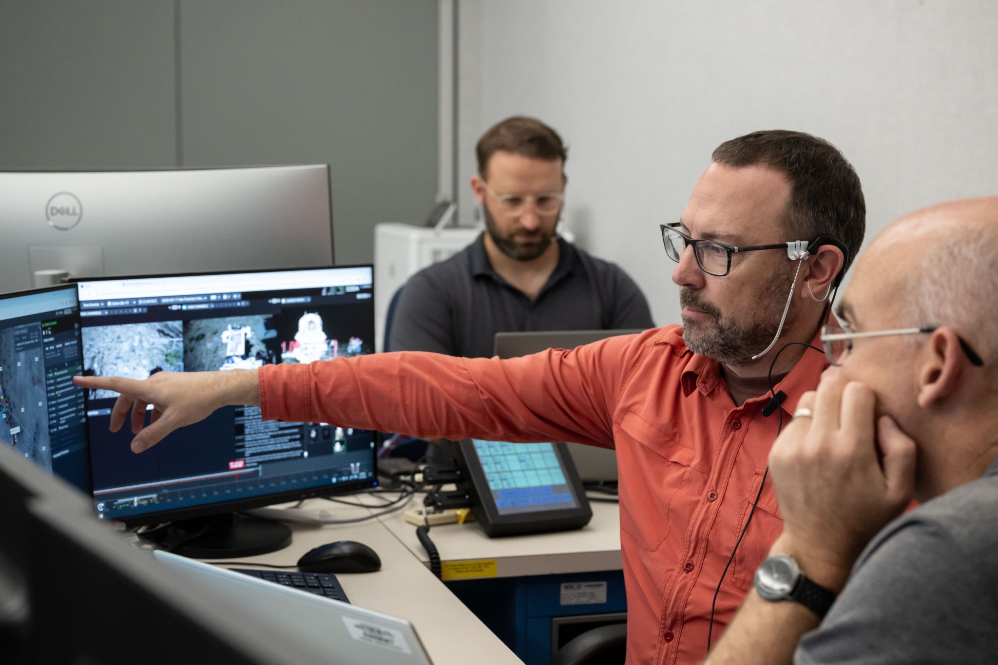 A person, wearing glasses, a headset, and a bright-colored shirt is in the center of the image, pointing at a computer screen — one of several visible in the image. In the background is another person, in glasses and a dark-colored polo shirt, looking down at this laptop screen. In the bottom right corner of the image a third person, wearing glasses, is visible from the side. That person is resting his head on his left fist, looking in the direction of the pointing hand. 