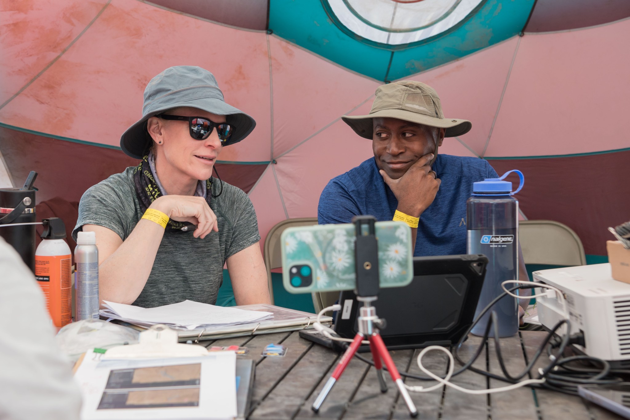 Two people sit side-by-side at a table inside a large tent. They’re wearing sun hats and t-shirts. The person on the left is talking and holding a pen in their left hand, while the person on the right is looking at them sideways and smiling. On the table in front of the pair is a jumble of papers, wires, an iPad and mobile phone propped up on stands, and large water bottles.